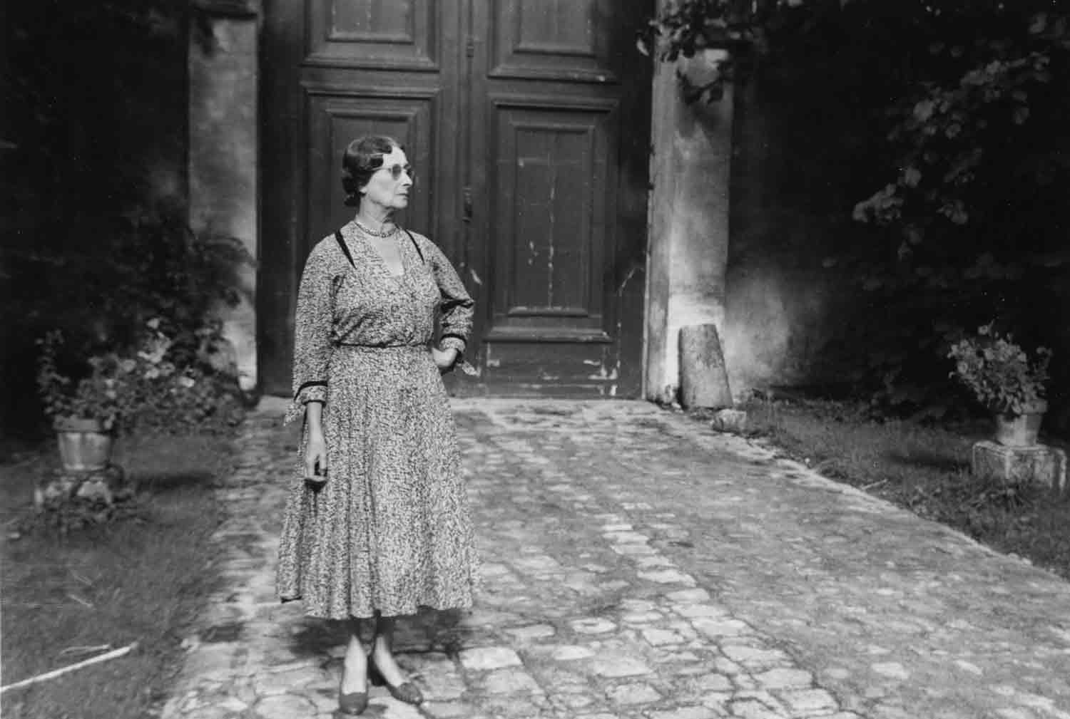 Black-and-white photo of a woman in a patterned dress and glasses, standing in a courtyard.