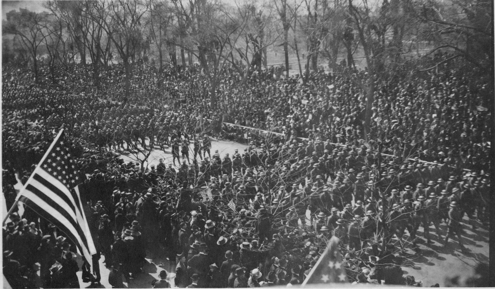 Soldiers marching in neat rows along Fifth Avenue in front of Central Park. An American flag hangs at left.