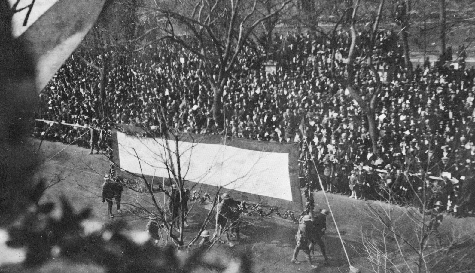 Soldiers carrying a large white banner in a military parade. The border and the letter H from another banner appear in the upper-left corner.