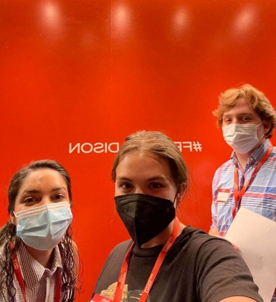 Three people in face masks posing for a selfie in front of a bright orange wall labeled with the hashtag #FrickMadison