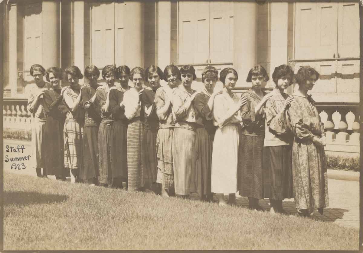 Archival photograph of fifteen women standing in a line in front of a building.