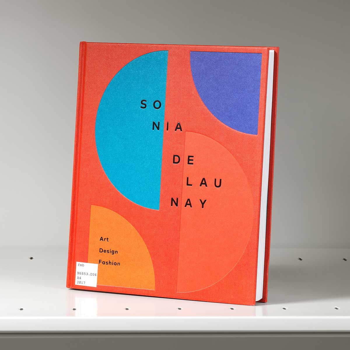 Book on a shelf whose cover features orange, red, and light and dark blue sections of a circle against a red background