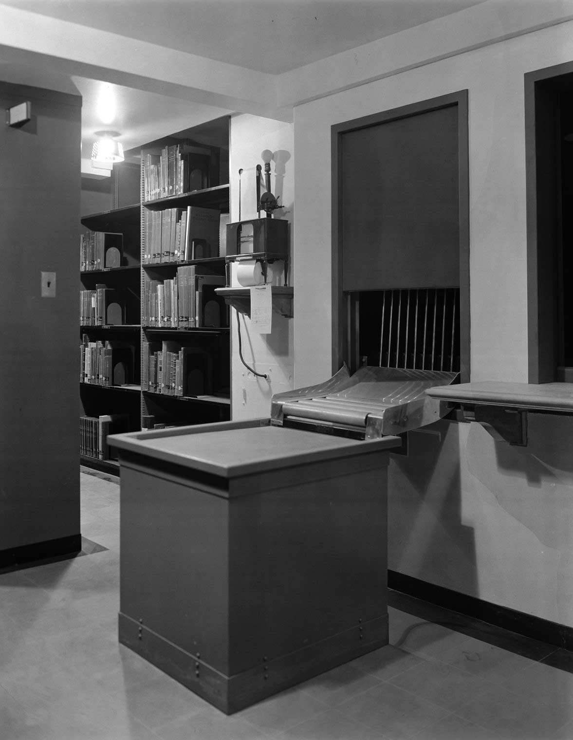 A station next to library book stacks with a desk, a telautograph machine, and a dumbwaiter