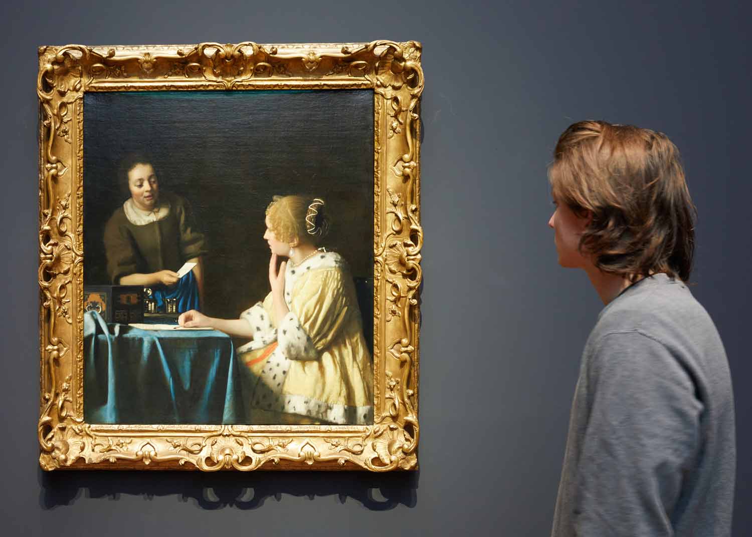 Man looking at a gold-framed painting of a maidservant handing a letter to a seated woman in a fur-trimmed yellow coat