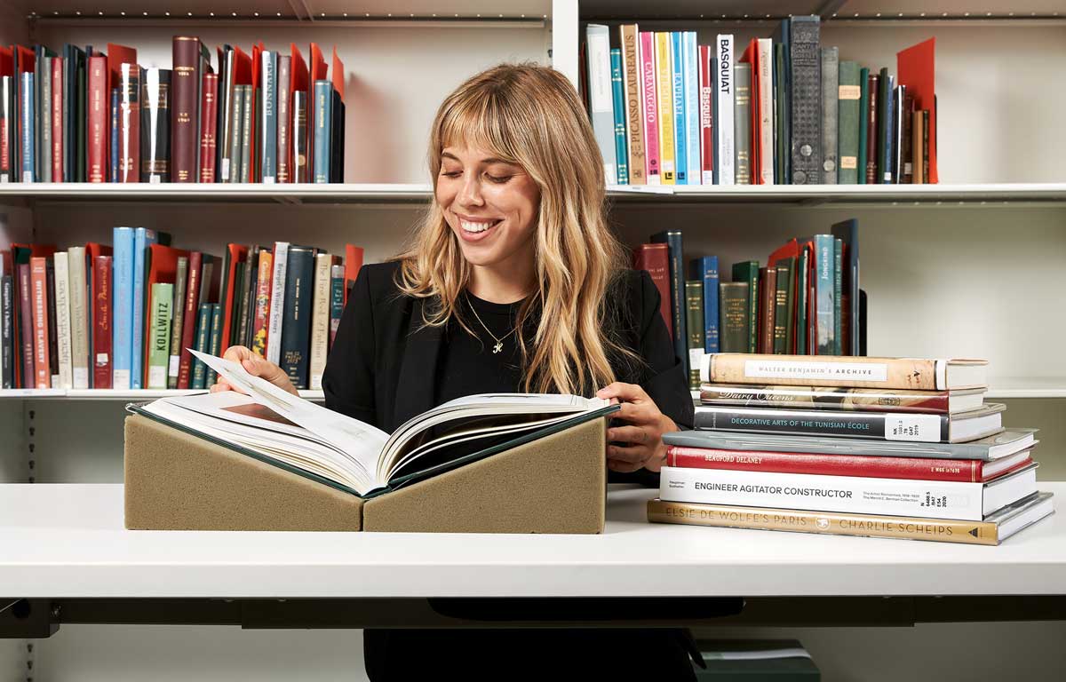 A blonde woman sitting at a desk leafing through a large volume next to a stack of books