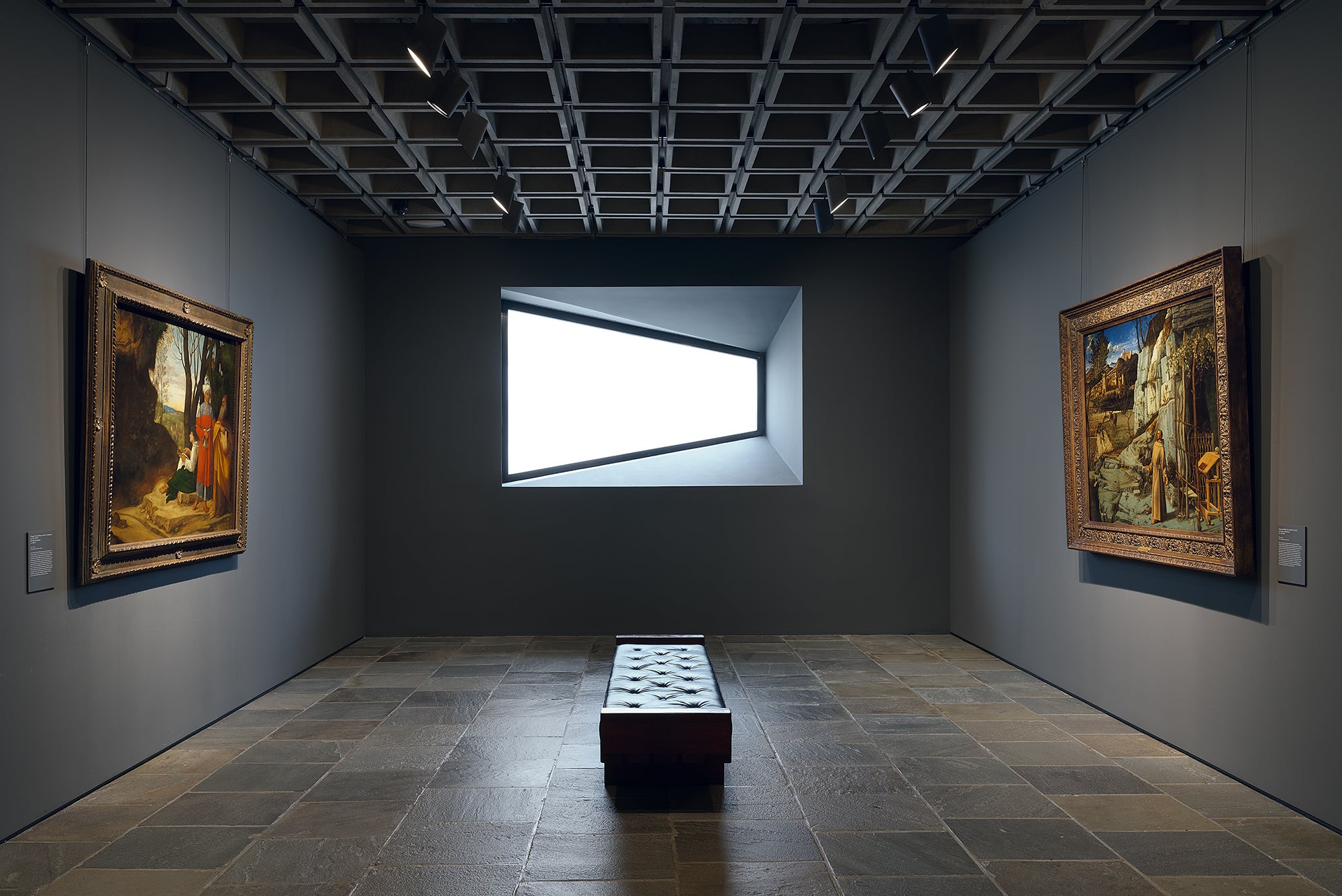 Gallery with two paintings hanging on opposite walls with a bench and trapezoidal window in between them