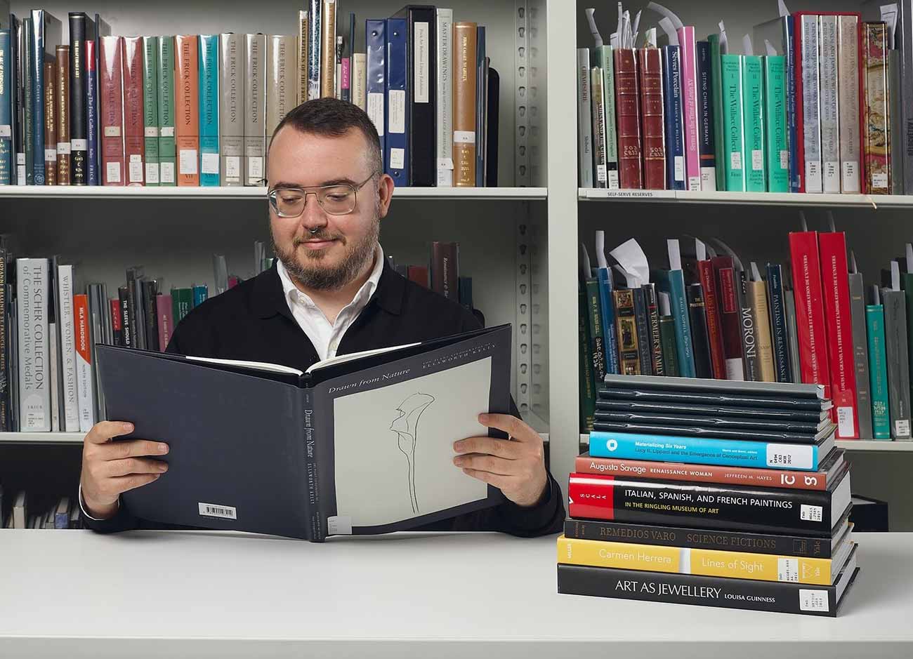 Man in a library flipping through a book at a desk stacked with 10 other books