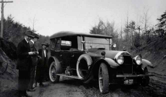 Black-and-white photograph of three people standing next to a car stopped in the middle of a dirt road.