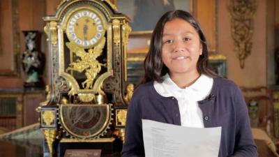 video still of young student standing next to clock on table