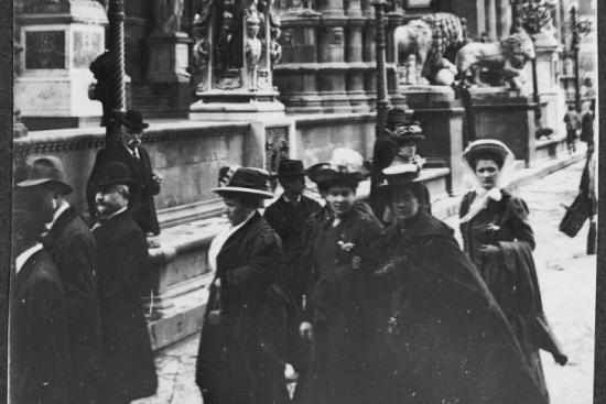 black and white photograph of a group of men and women wearing cloaks and hats walking in front of stone columns and sculptures 