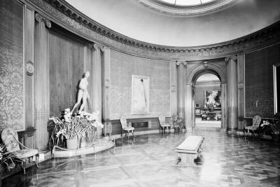 photo of oval shaped gallery room with sculpture, plants chairs, paintings, bench, circa 1976