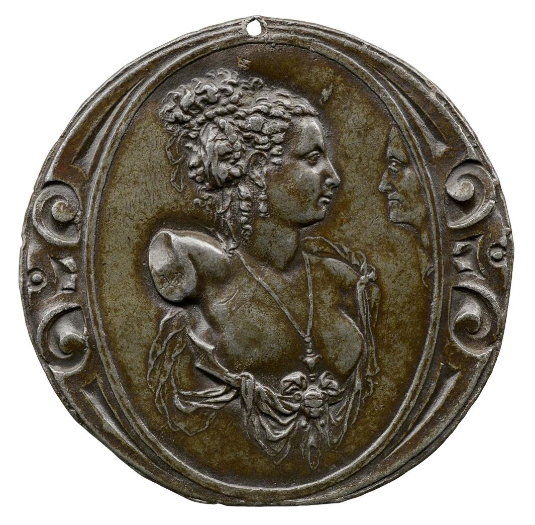 Lead medal of a bust of a nude woman surrounded by drapery, wearing a necklace and earrings and ribbons in her curly hair, in profile to the right with a face in profile looking in from the right