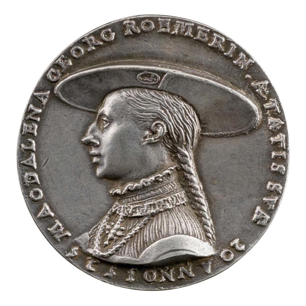 Silver medal of a man with long hair pulled back in braid, wearing a wide hat with badge