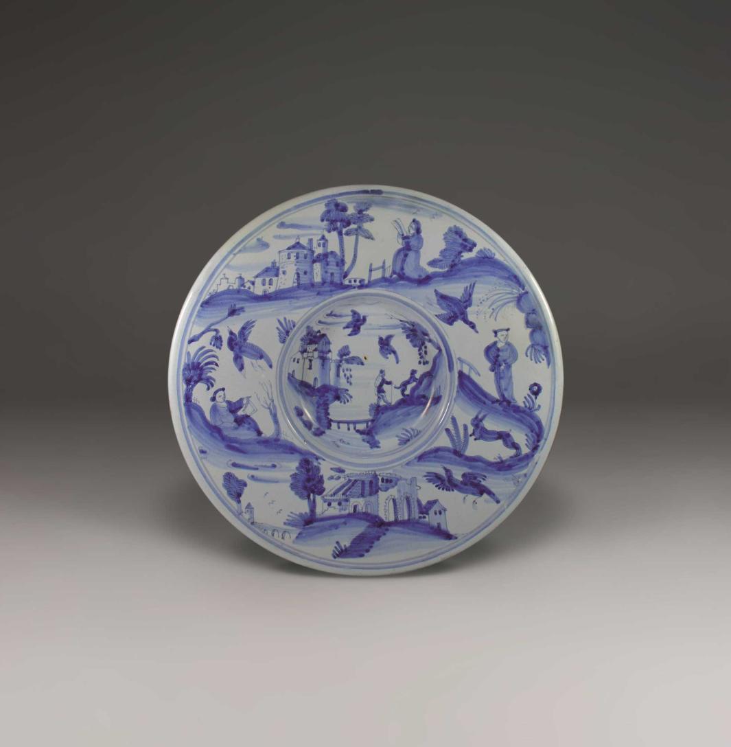 Earthenware plate in blue and white with a landscape scene of a figure and dog walking uphill at the center and more landscape scenes of figures and animals on the outside.