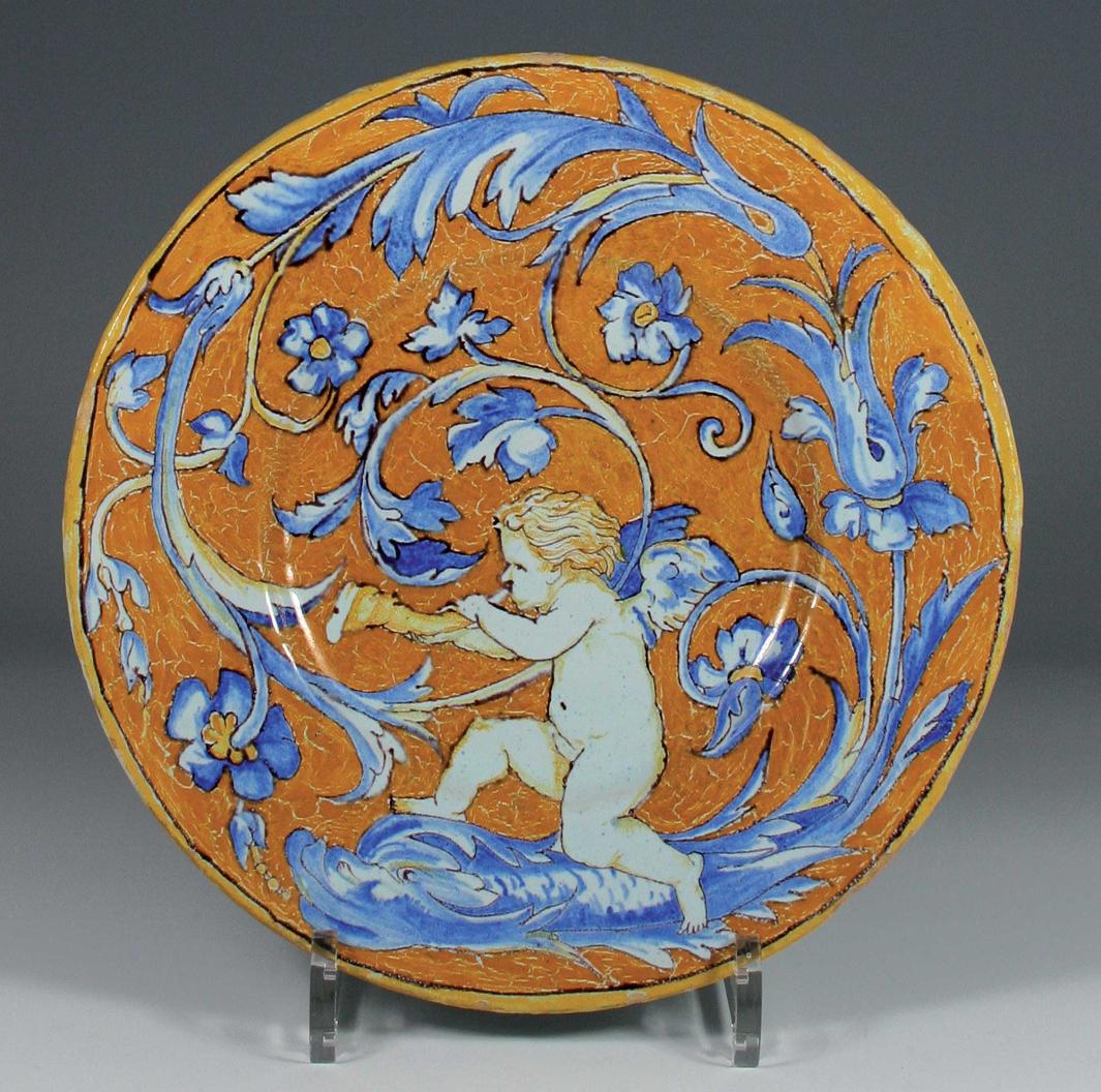 Earthenware plate with a winged nude figure blowing a horn and sitting on a fish, whose tail turns into a floral design swirling around the plate.