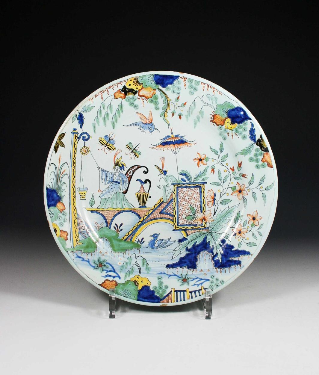 Earthenware plate with a landscape scene of a figure sitting on a bridge using a pulley system to pull a bucket of water from the stream below and another figure watching.