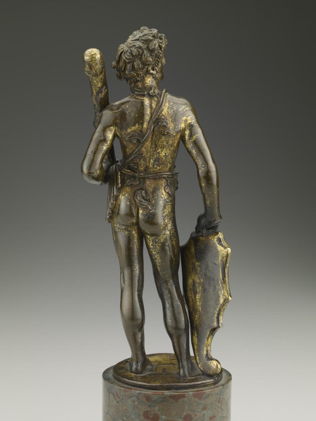 Back view of a bronze sculpture of a man standing upright.  His head is turned to his right and he is holding a club in his left hand.  A shield sits by his right leg and is underneath his right hand.