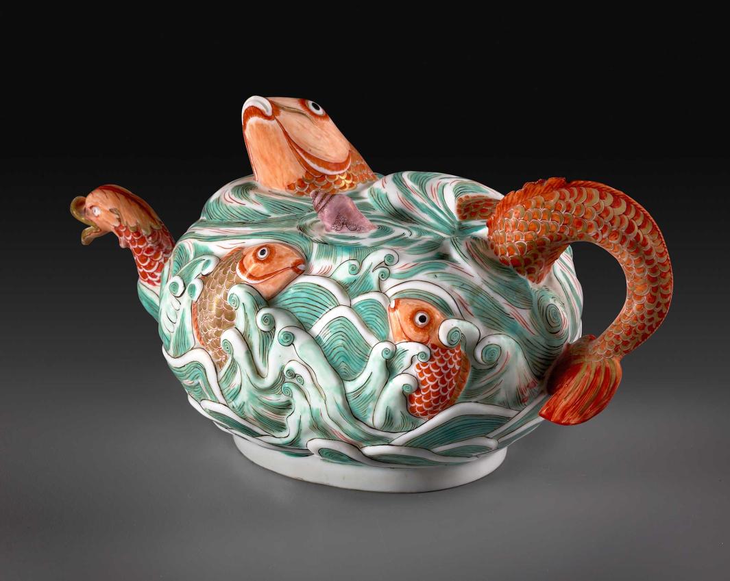 Three-quarter view of porcelain teapot depicting high relief fish in water in orange and green glaze