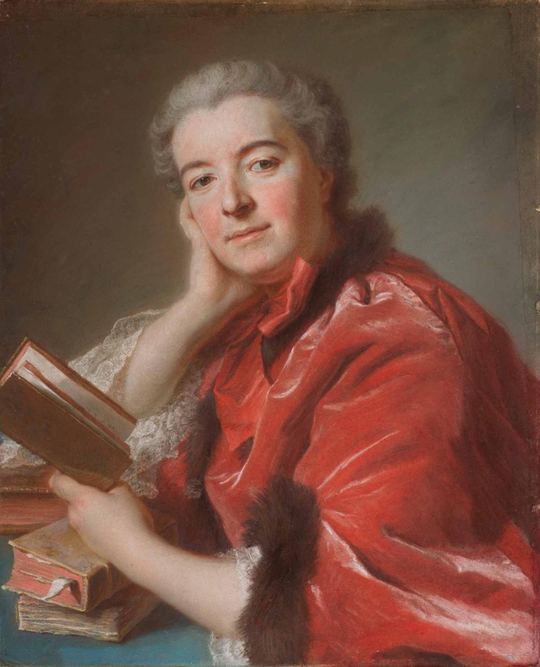 *A gray-haired woman in a pink coat with brown fur trim leans on a table with books on it, holding a book in her left hand while she looks out at the viewer, her right hand resting on her right cheek*