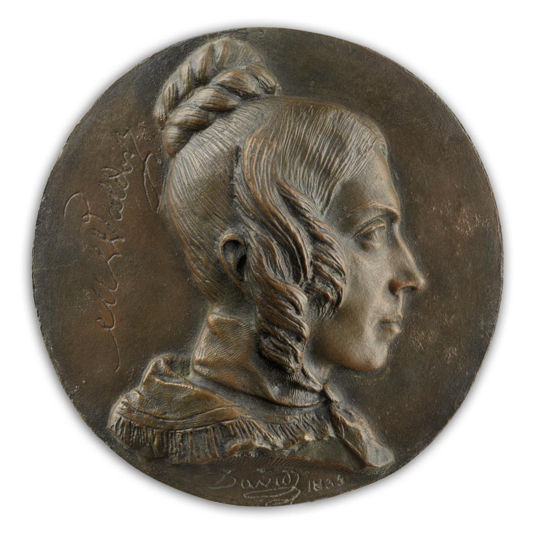 bronze head of woman in profile with hair in bun and side locks, atop circle of bronze with writing