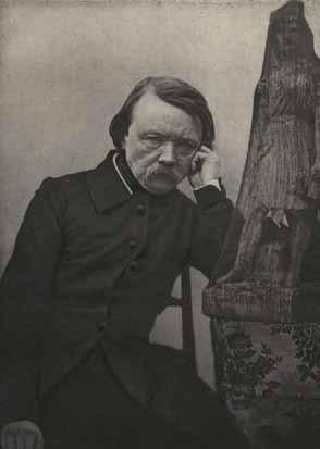 black and white photograph of middle-aged man with moustache dressed in black, leaning one elbow on a sculpture