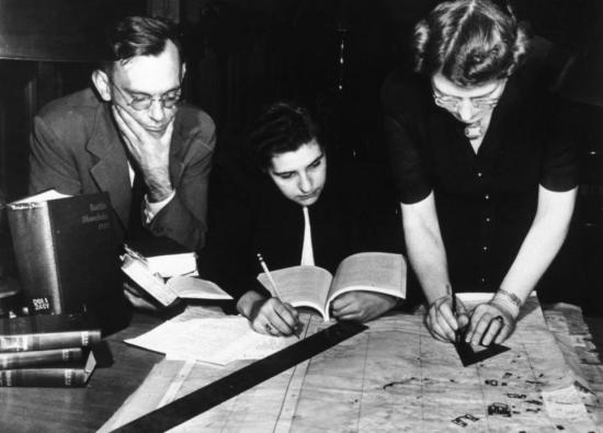 Three individuals at a desk, surrounded by books and studying a map that they are marking with rulers and pens