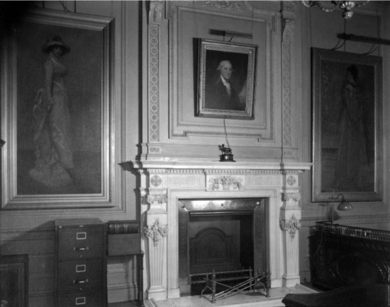 Black-and-white photo of an office wall in a Gilded Age mansion. A portrait of George Washington hangs over a fireplace flanked by two portraits of fashionable women.