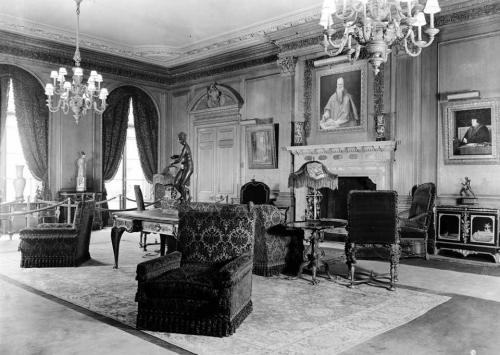 Black-and-white photo of The Frick Collection's Living Hall gallery, with furniture, a statue of a woman, two portraits flanking a fireplace, and a larger portrait above the mantelpiece