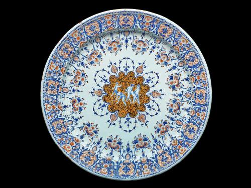 Earthenware platter with four nude figures in the middle surrounded by floral designs in blue and orange