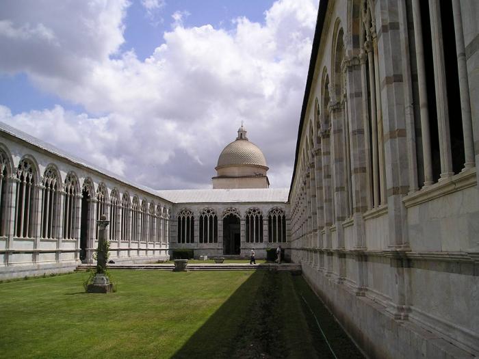 Colonnaded walkways of a medieval building surrounding a grassy courtyard, with a dome in the distance