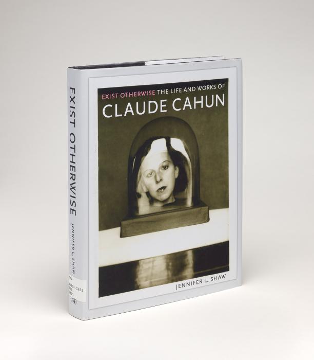 Cover of "Exist Otherwise: The Life and Works of Claude Cahun" against a gray background, featuring a photograph of Cahun's face in a bell jar on a table reflecting light