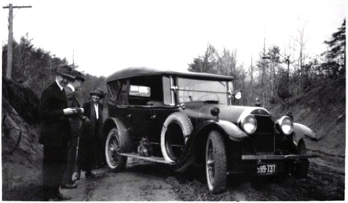 Black-and-white photograph of a woman and two men standing next to an early automobile parked on a muddy dirt road.