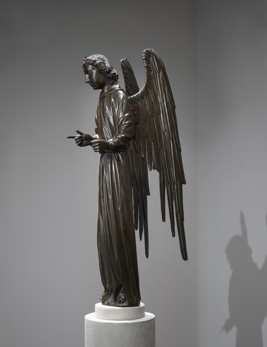 Installation view of a bronze sculpture of an angel. The angel stands upright with wings stretched behind it. The right pointer finger points out and the angel glances downward.