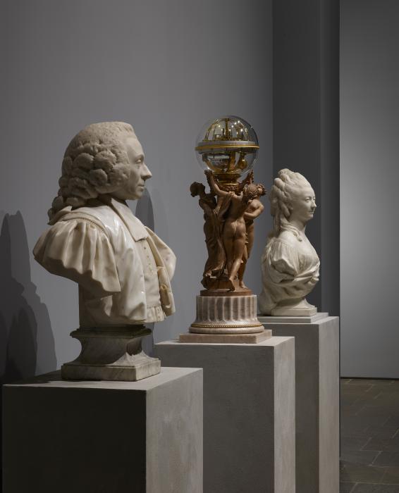 Installation view of a clock encased in a glass globe held aloft by three terracotta female figures. The clock sits between two marble sculptures of a man, at left, and a woman, at right, both with elaborate hairstyles.