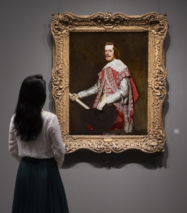 Gallery view of an oil painting in a gilt frame of King Philip IV of Spain dressed in a silver and rose costume and holding a black felt hat and ivory-colored baton. A woman stands in front of the painting, her back to the viewer.