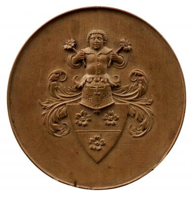 Wooden model for a medal depicting the coat of arms of Matthaeus Schwarz which consist of three flowers on a shield, surmounted by a helmet and foliage, on top of which is the torso and head of a small man holding flowers in both hands