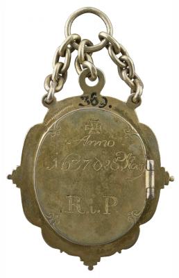 Back of a gilt case for a medal suspended from a ring by three short chains, with a hinge at right and an inscription on the case.