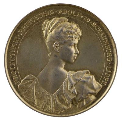 Copper medal of a woman overlooking her right shoulder wearing a dress with a low back and lace collar