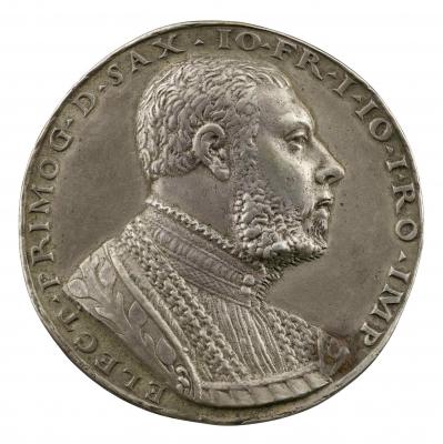 Silver medal of a man wearing a pleated shirt, several chains around his neck and an embroidered mantel