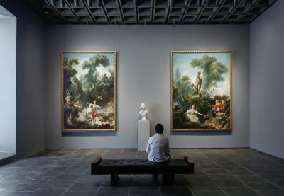 Man seated in a gallery with two large, colorful Rococo paintings and a marble bust