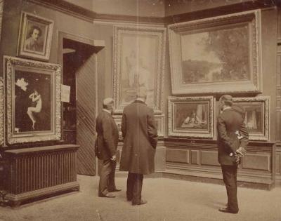 Three men in long coats viewing framed paintings hanging on a wall