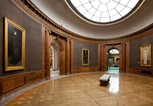 color photograph of oval gallery with grey walls, wooden pillars, polished floor, and skylight
