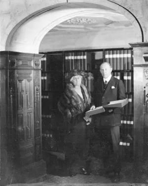 Lady and Sir Robert Witt standing in front of library stacks with binders, circa 1923