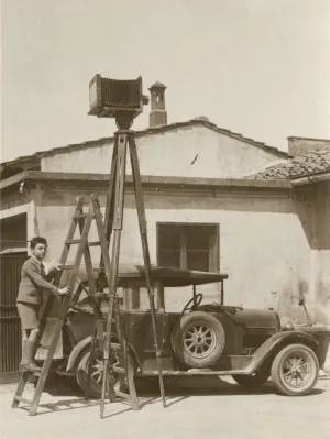 young boy at foot of ladder, next to elevated camera, next to car and small house, circa 1938