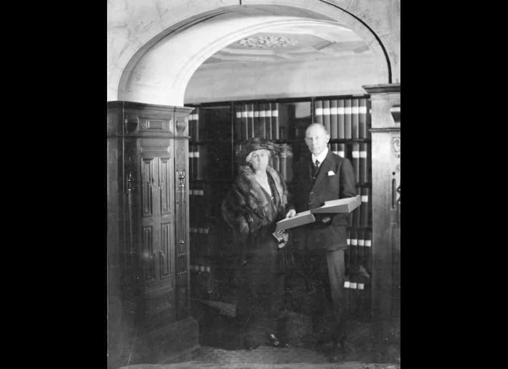 Lady and Sir Robert Witt standing in front of library stacks with binders, circa 1923